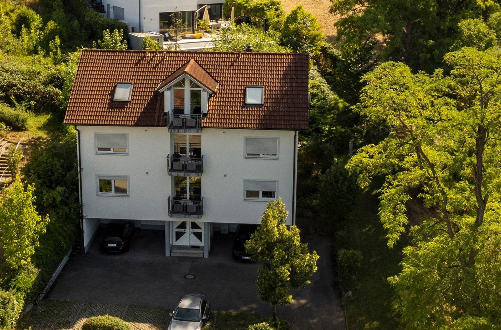 Das Gebäude von oben: weiß verputztes Mehrfamilienhaus mit rotem Ziegeldach, drei übereinanderliegenden Balkonen und überdachten Stellplätzen unter dem Haus, umgeben von Bäumen