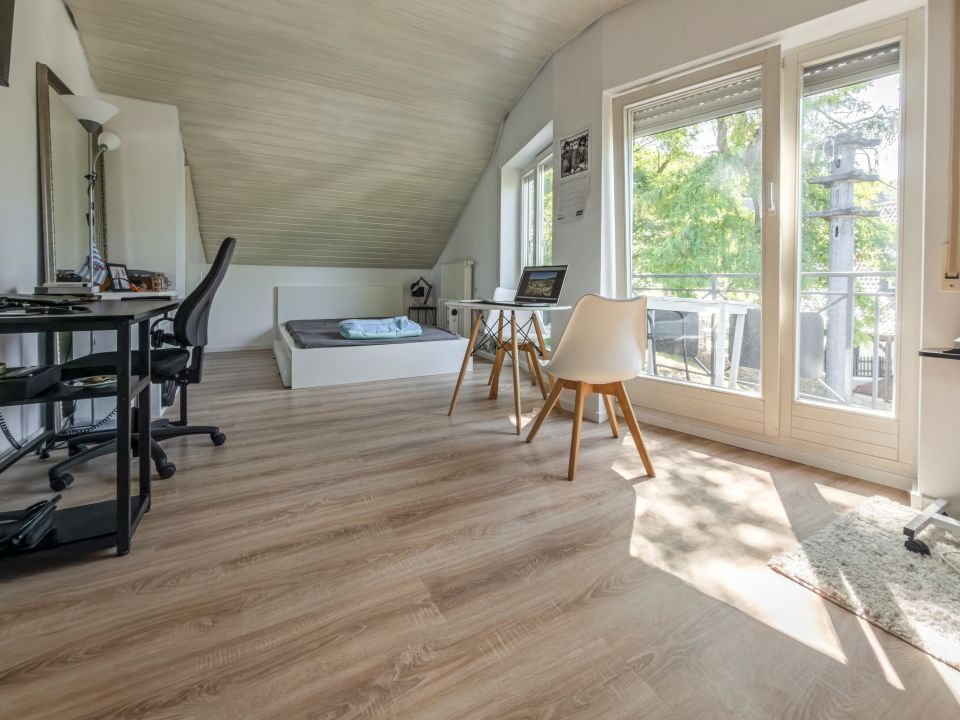 Quartier im Dachgeschoss mit Dachschräge, bodentiefem Fenster zum Balkon, Bett und zwei Arbeitsplätzen